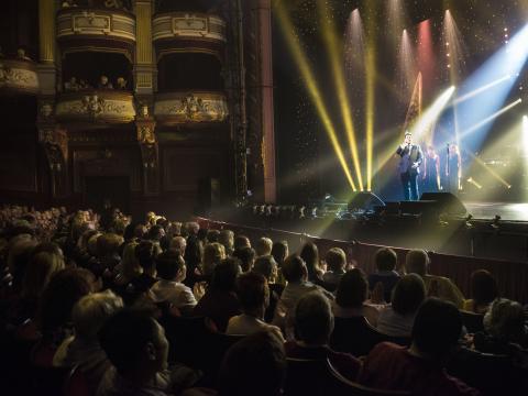 Audience sat watching Michael Ball performing on stage at the Alhambra Theatre with yellow stage lights shining upward.